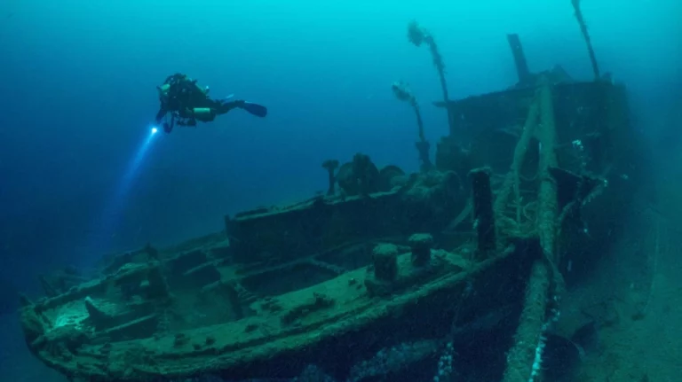A scuba diver swimming over a wreck in a drysuit holding a torch