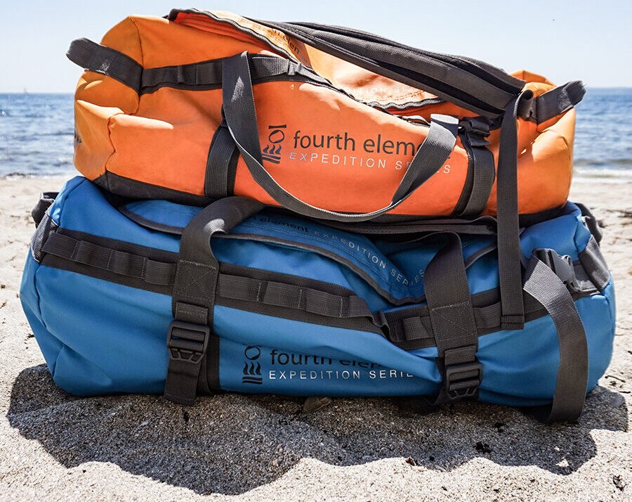 Fourth Element Expedition Series Duffel Bags in blue and orange stacked on beach